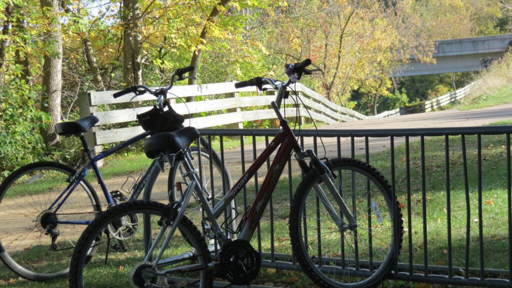 Two bicycles parked by a trailhead with a white fence and trees in fall colors.
