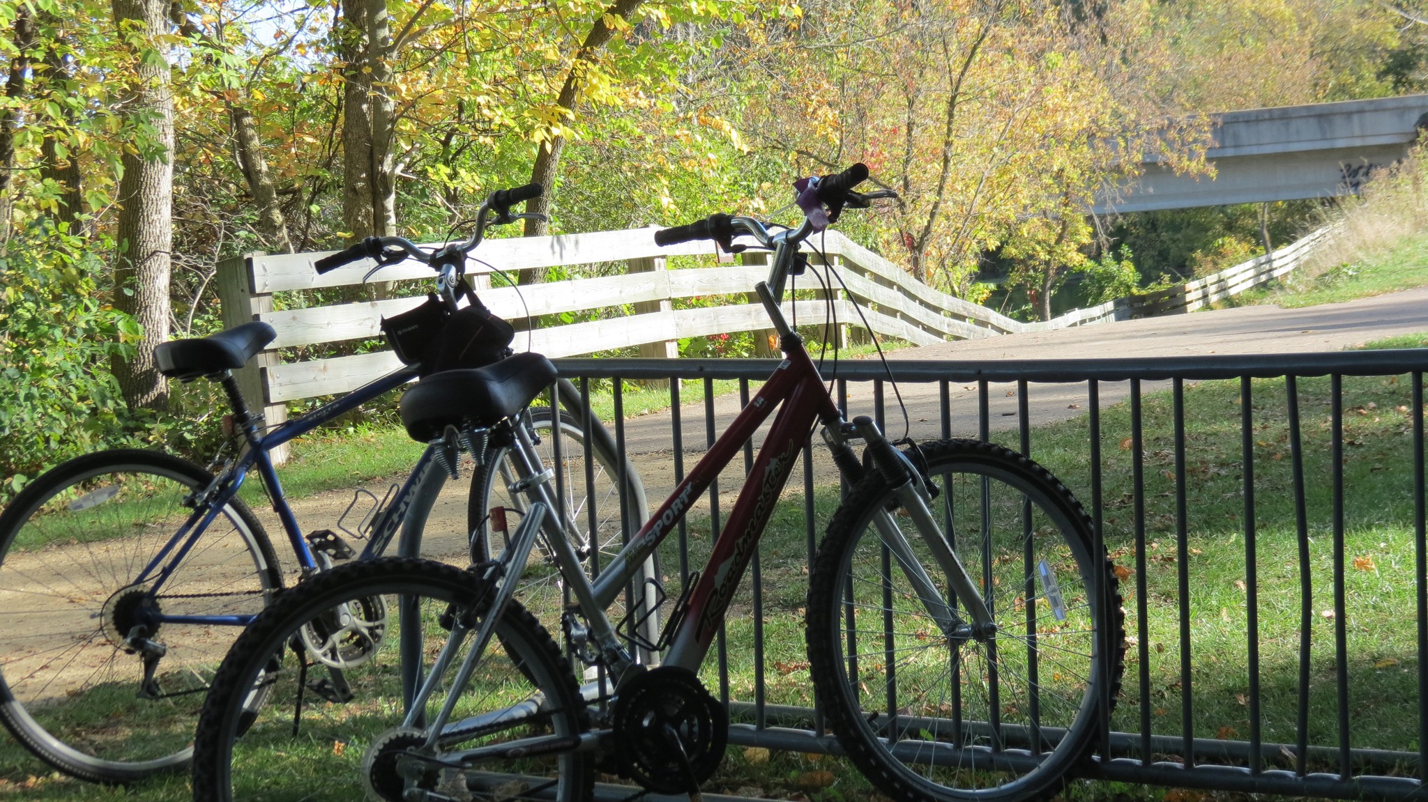 Two bicycles parked by a trailhead with a white fence and trees in fall colors.